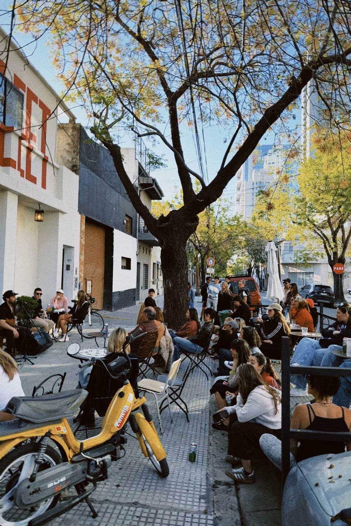 La vereda del club llena de gente escuchando un show en vivo, bajo el árbol